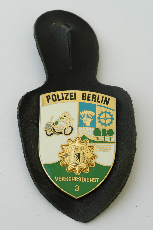Ein Polizeistern mit der Aufschrift "Polizei Berlin", der ein blaues Schild mit einem weißen Stern in der Mitte zeigt, umgeben von einem weißen Rand, auf einer Oberfläche platziert.