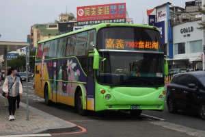 Grüner Bus fährt auf einer Straße mit hohen Gebäuden, eine Frau geht auf dem Gehweg und trägt eine Tasche, Bäume, Pfosten, Plakate und einen klaren blauen Himmel.