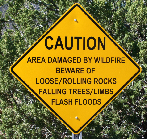 Gelbes Warnschild an einem Pfosten im Vordergrund, das vor Wildfire-Schäden, lockeren Steinen, umstürzenden Bäumen und Überschwemmungen warnt, mit Bäumen im Hintergrund.