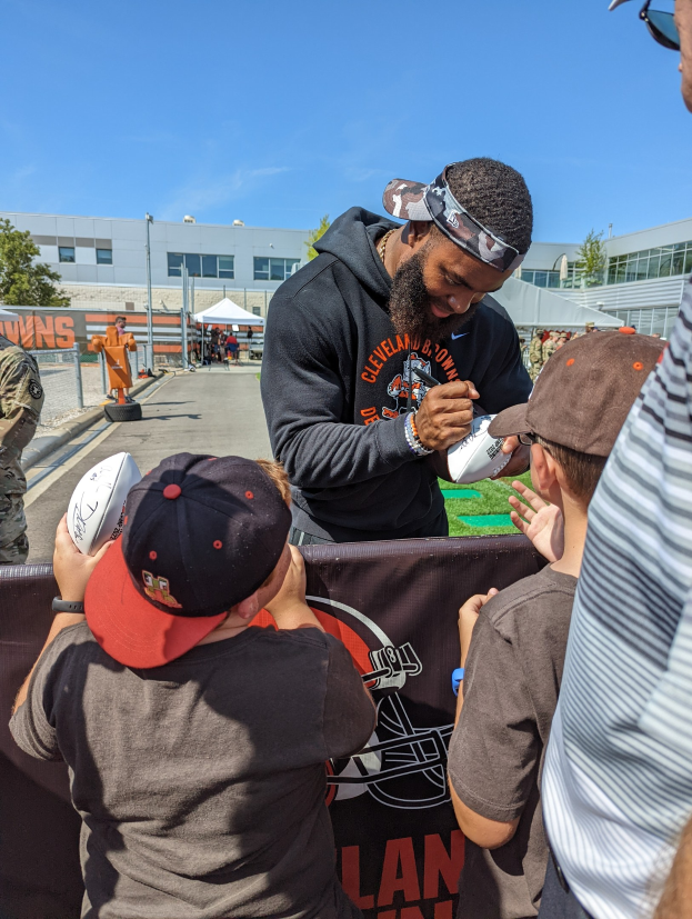 Cleveland Browns Wide Receiver Odell Beckham Jr. signiert Autogramme für Fans mit einer Mütze und Schutzbrille auf dem Trainingsgelände des Teams, umgeben von Menschen mit Mützen, einem Banner und Gebäuden im Hintergrund.