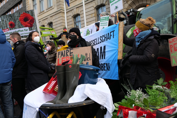 Maskierte Demonstranten halten Schilder vor einem Lastwagen, mit einem Tisch voller Gegenstände, Topfpflanzen und einem Gebäude mit Flagge im Hintergrund, gegen ein Pestizidverbot.