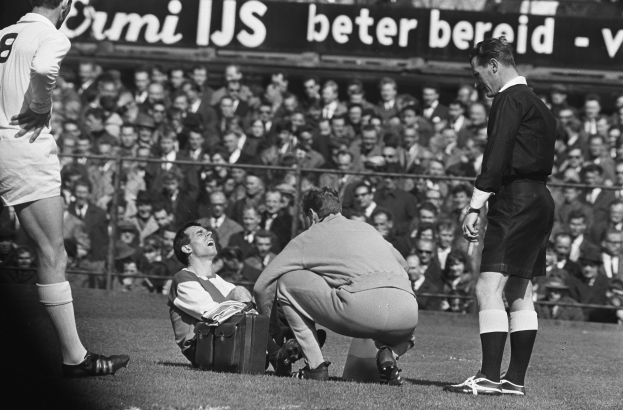 Ein Schwarz-Weiß-Foto eines Schiedsrichters, der mit einem Mann auf dem Feld spricht und von einer Gruppe Menschen umgeben ist, mit einem Zaun, einer Fahne und Bäumen im Hintergrund.
