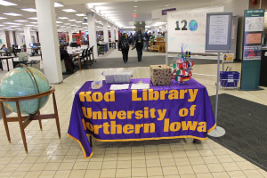 Eine Ausstellung mit einem Tisch, der verschiedene Landesflaggen, eine Box, Papiere und eine Fahne zeigt, eine Kugel links daneben, ein Anschlagbrett rechts daneben mit ein paar Leuten in der Nähe und mehreren Tischen und Stühlen im Hintergrund.
