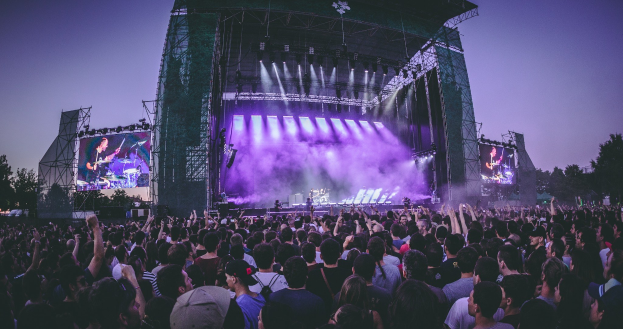 Eine große Menschenmenge auf einem Musikfestival schaut auf einer hell erleuchteten Bühne mit Instrumenten und Bildschirmen, die vor einem Gebäude, Bäumen und einem klaren blauen Himmel steht.