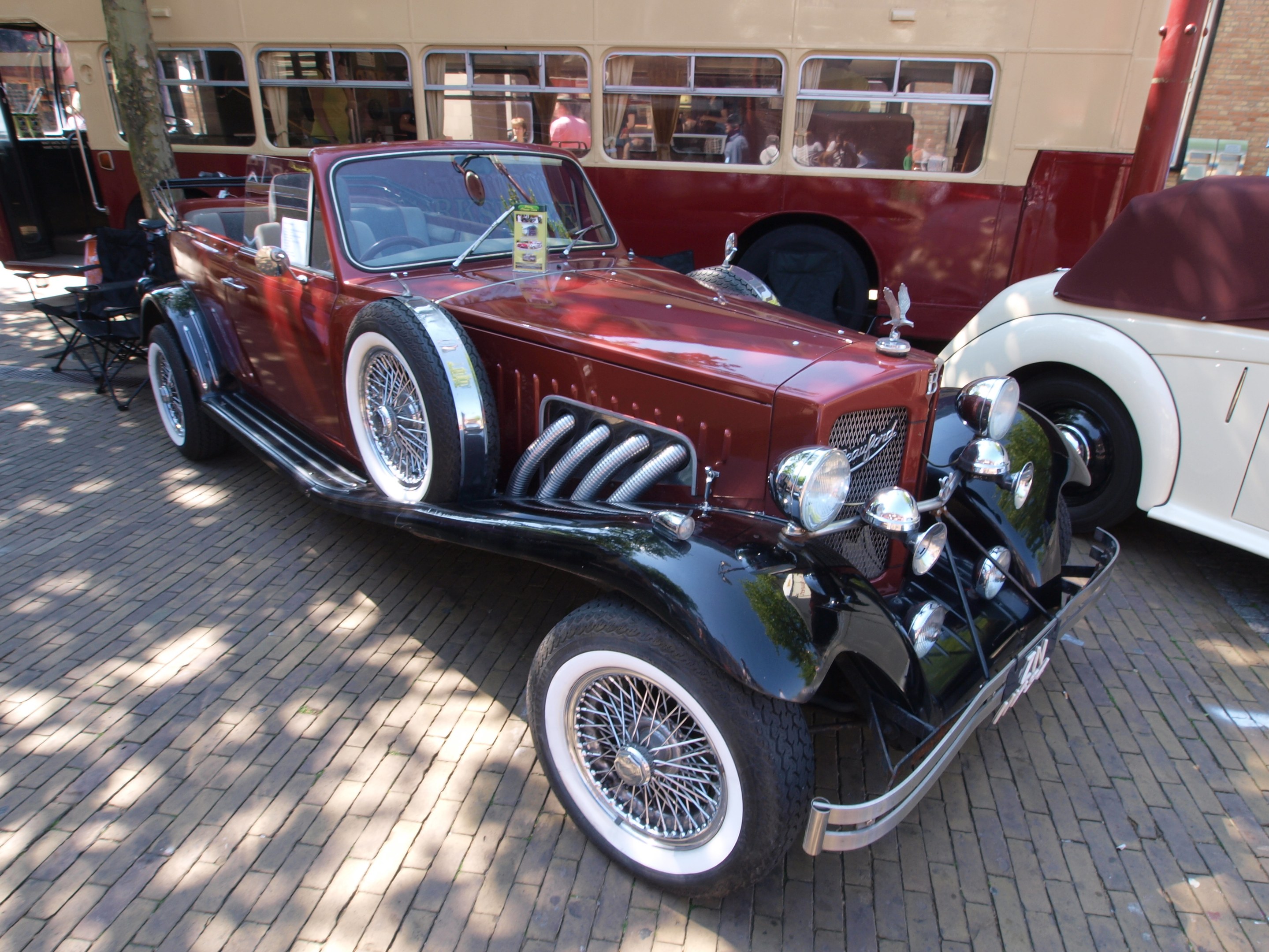 Ein Vintage-Auto neben einem Doppeldeckerbus auf einer Kopfsteinpflasterstraße geparkt, mit Stühlen und einem Baum im Vordergrund, einem Gebäude mit Fenstern im Hintergrund und Menschen im Bus zu sehen.