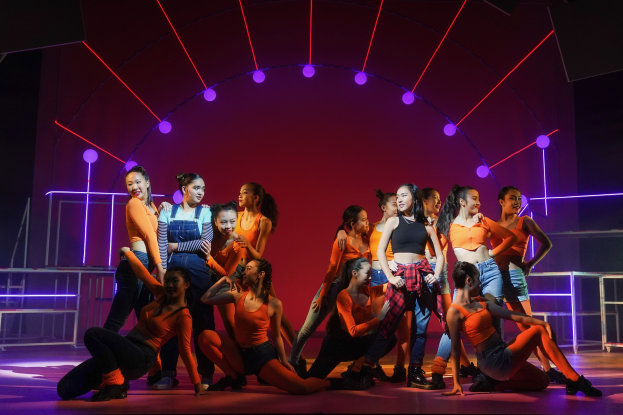 Eine Gruppe junger Tänzer in bunten Kostümen performs auf einer Bühne mit Geländern, beleuchtet von bunten Lichtern, mit einem jubelnden Publikum im Hintergrund.