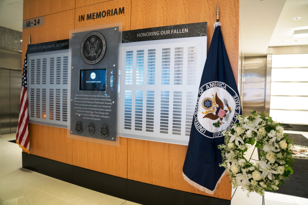 Gedenktafel in einer Lobby mit Flaggen, einem Kranz und einem Blumenstrauß, einer Wand mit Brettern und Text, beleuchteter Decke und Aufzügen auf der linken Seite.