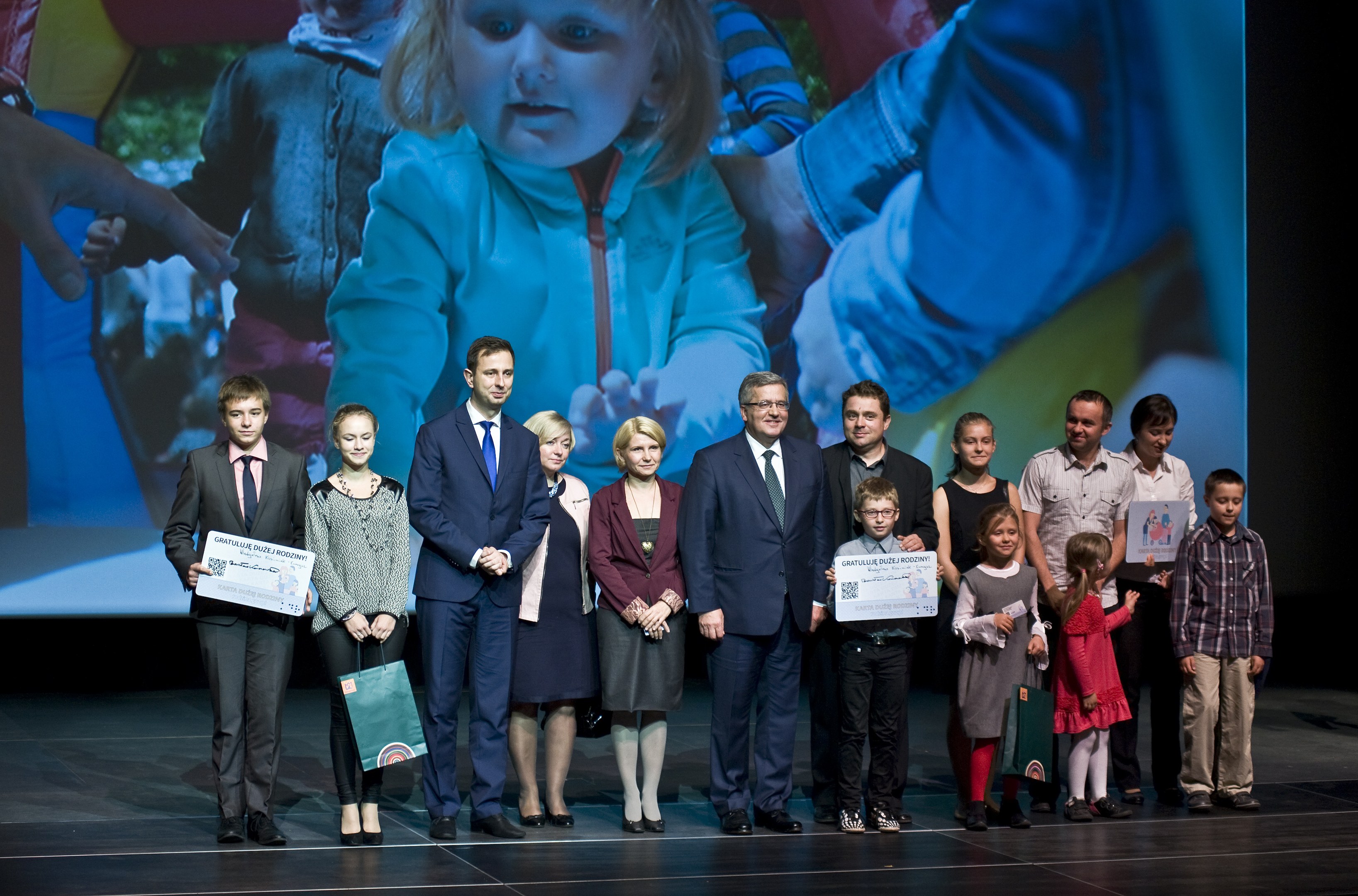 Eine Gruppe von Menschen auf einer Bühne mit Taschen und Urkunden, im Hintergrund ein Bildschirm mit einem Kind und anderen.