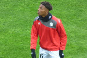 Ein junger Mann in einer roten Jacke und weißen Shorts steht auf einem Fußballfeld und schaut rechts mit entschlossener Miene, trägt Baseball-Handschuhe.