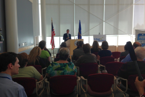 Ein Mann steht an einem Rednerpult und spricht zu einem Publikum, das auf Stühlen sitzt, mit zwei Fahnen daneben und einer Wand, Säulen und Fenstern im Hintergrund.