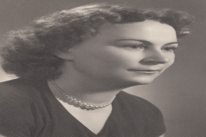 Ein Schwarz-Weiß-Foto einer Frau mit ernstem Gesichtsausdruck, die eine Kette und ein einfaches Kleid mit hohem Kragen und langen Ärmeln trägt.