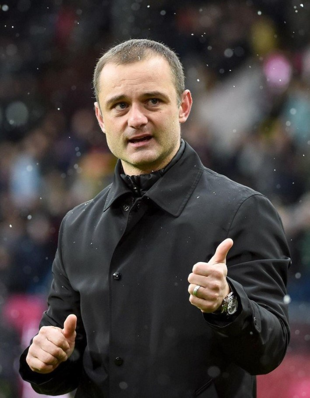 Ein Mann in einer schwarzen Jacke zeigt einen Daumen hoch und trägt eine Uhr, mit einem unscharfen Hintergrund.