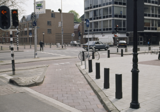 Eine Straßenszene in der Stadt mit einem geparkten Fahrrad, fahrenden Fahrzeugen, Fußgängern, Pfosten, Verkehrsampeln, Schildern, Bäumen, Gebäuden mit Fenstern und einem klaren blauen Himmel.