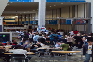 Ein belebter Flughafenvorplatz mit wartenden Menschen, einige stehen und andere arbeiten, Richtungsschilder an der rechten Wand und an der Decke sowie Deckenleuchten.