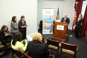 Drei Frauen sitzen auf Stühlen, während ein Mann und eine Frau an einem Rednerpult in der Mitte stehen, mit zwei Fahnen und einem Banner im Hintergrund; links sieht eine Frau auf eine Digitalkamera, während zwei andere Frauen stehen, und Vorhänge sind in der oberen rechten Ecke sichtbar.