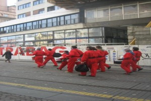 Menschen, die auf einem Boden gehen und Musikinstrumente spielen, mit einer Wand aus Gittern, die Plakate und Text trägt, und weiter hinten Gebäude mit Glaswänden und Fenstern, mit Namen auf den Gebäuden.
