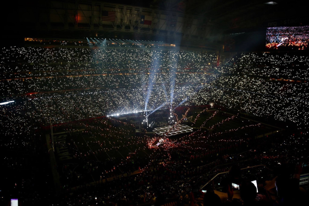 Eine große Menschenmenge in einem hell erleuchteten Stadion bei Nacht schaut auf einen Bildschirm, der die Ergebnisse der Super Bowl XLVII-Halbzeitshow anzeigt.