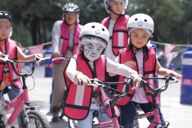 Gruppe junger Mädchen, die Fahrräder auf einer Straße fahren, tragen Helme und Schwimmwesten, mit Bäumen und unscharfem Hintergrund.