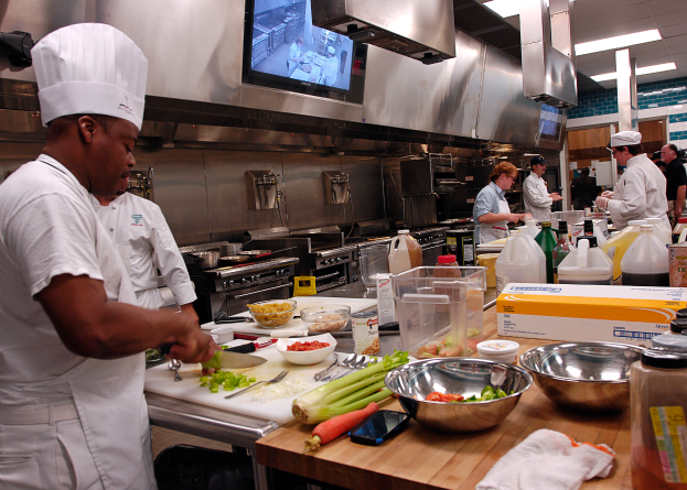 Eine Gruppe von Köchen in einer Küche beim Zubereiten von Essen, mit einem Koch im Vordergrund in einer weißen Schürze und Mütze, der ein Messer hält und von verschiedenen Utensilien, Schüsseln, Gemüse, einem Handy und Flaschen umgeben ist, mit einem Fernseher und Deckenlampen im Hintergrund.