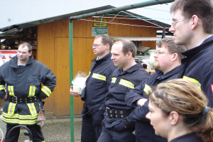 Eine Gruppe von Männern in blauen Feuerrettungsuniformen steht und schaut nach links, mit einem kleinen hölzernen Schuppen und Rohrrahmen im Hintergrund.