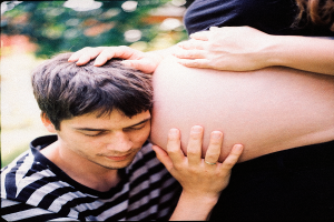 Ein Mann und eine Frau stehen nebeneinander, der Mann berührt zärtlich den Bauch der Frau, mit einem unscharfen Hintergrund, der das Paar und ihr ungeborenes Kind hervorhebt.