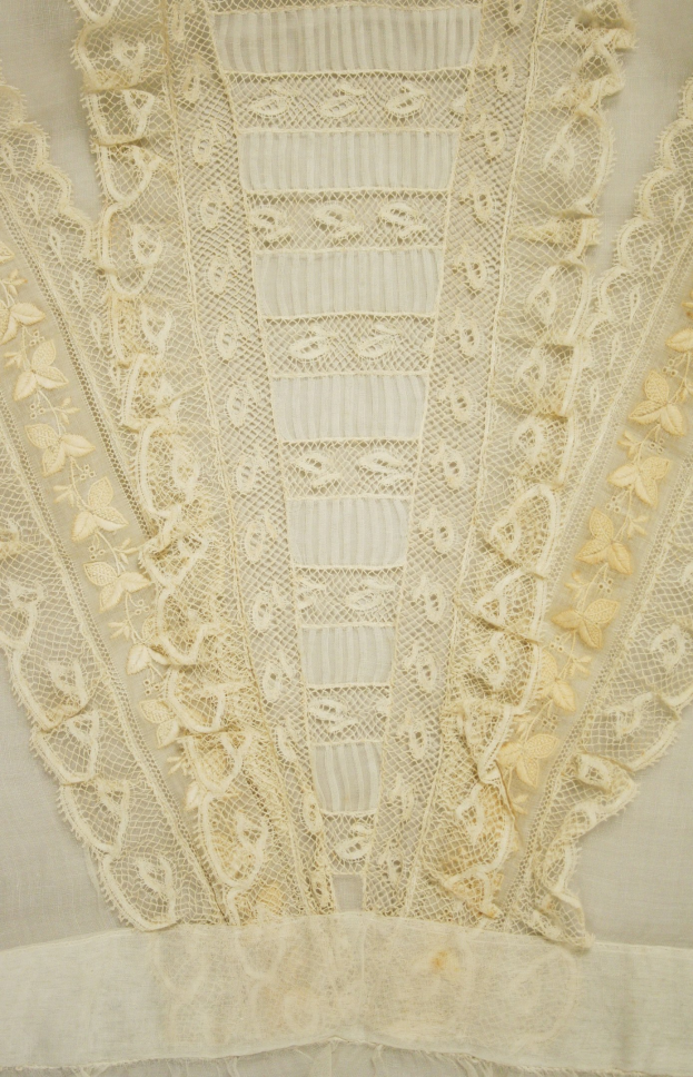 Nahaufnahme eines weißen Kleides mit zarter, detailreicher Spitzenverzierung mit verschiedenen Formen und Mustern auf leichtem, luftigem Stoff.
