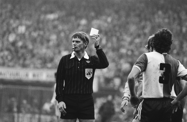 Schwarzes und weißes Bild eines Schiedsrichters, der eine rote Karte vor einer Menge von Menschen mit Fußballtrikots hält, wobei ein Zaun im Hintergrund zu sehen ist.