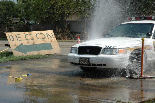 Ein Polizeiauto fährt durch eine Pfütze, mit einer Tafel, einem Verkehrskegel und anderen Gegenständen auf dem Boden, mit Bäumen, Gebäuden und einem klaren blauen Himmel im Hintergrund.