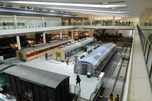 Ein großer Museumsraum mit verschiedenen Zügen in unterschiedlichen Größen und Farben, mit Menschen, die die Ausstellungsstücke beobachten und Deckenleuchten, die den Raum erhellen.