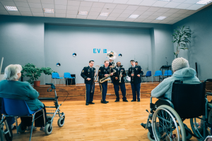 Eine Gruppe von Menschen im Rollstuhl, die in einem Raum mit Stühlen, Pflanzen und Deckenleuchten Musikinstrumente spielen, mit ein paar Menschen auf Stühlen, was auf ein Rollstuhlband-Event hindeutet.