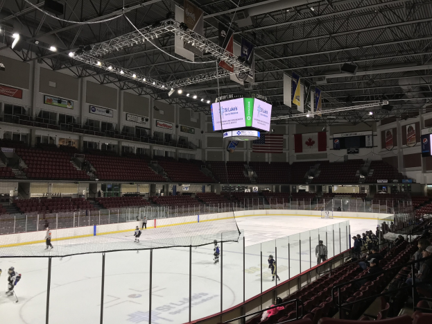 Hockeyspiel in einer großen Arena mit einem zentralen Netz, Zuschauern auf Stühlen, Deckenbeleuchtung und Trägern sowie Bannern an den Wänden, identifiziert als die St. Louis Blues Arena.