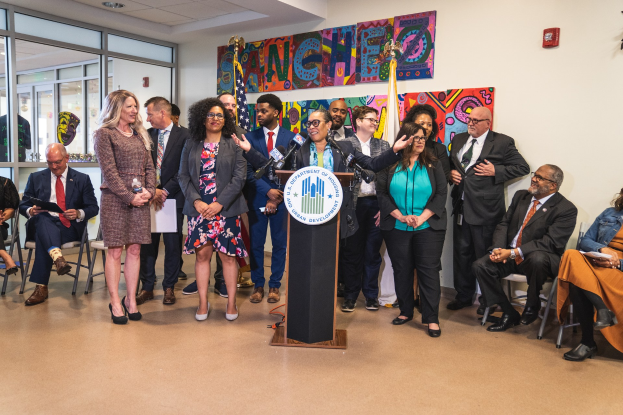 Gruppe von Menschen vor einem Rednerpult mit Mikrofon und einer Tafel mit Text, einige sitzen auf Stühlen und andere stehen, mit Fahnen, einer Tafel, einem Feuerlöscher und Deckenleuchten an der Rückwand.