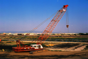 Ein Baukran steht auf einem freien Feld mit verstreuter Ausrüstung, während Häuser, Gebäude, Bäume, Pflanzen und Berge im Hintergrund zu sehen sind.