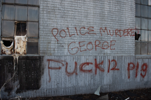 Ein Gebäude mit Graffiti, das 'Polizei Mord George Fuck 12 P19' auf der Seite trägt, mit Fenstern und verschiedenen Gegenständen vorne.