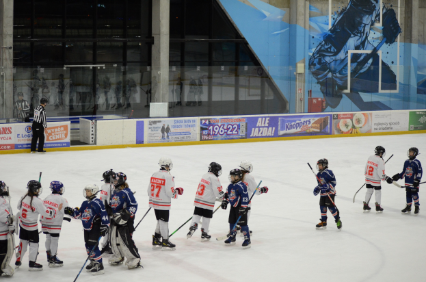 Gruppe junger Menschen, die Eisockey auf einer Indoor-Eisfläche spielen, mit Helmen und Stöcken, sowie Bannern, Säulen und Glaswänden im Hintergrund.