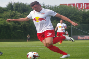 Ein Mann in einem Fussballtrikot schie├čt einen Ball auf einem gr├╝nen Feld mit B├Ąumen und einem klaren blauen Himmel im Hintergrund, mit einem Schild mit der Aufschrift "RB Leipzig".