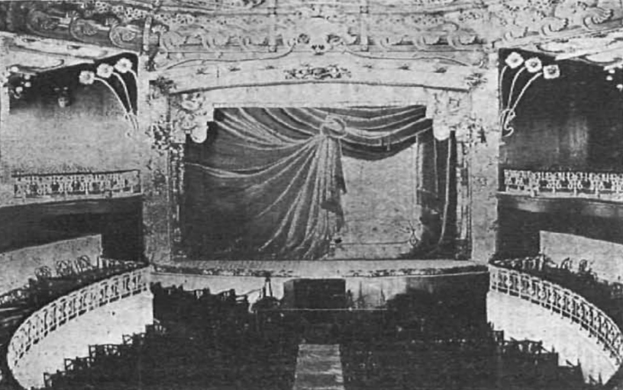 Schwarzes und weißes Foto des Theatersaals des Theatre Royal in London, das eine Bühne mit Vorhang, Geländern und verzierte Wanddesigns zeigt, mit einigen sitzenden Menschen und Text unten.