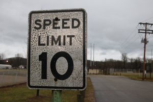 Geschwindigkeitsbegrenzungsschild am Rande einer Straße mit umliegender Wiese, Strommästen, einem Zaun, Häusern, Bäumen und einem bewölkten Himmel im Hintergrund.