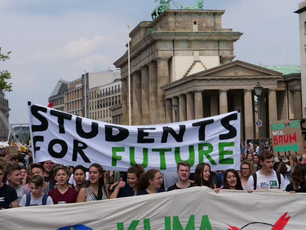 Eine Gruppe von Schülern marschiert in Berlin, hält eine bunt bemalte 'Students for Future'-Schultertasche gegen eine Kulisse aus Gebäuden, Bäumen und Himmel.