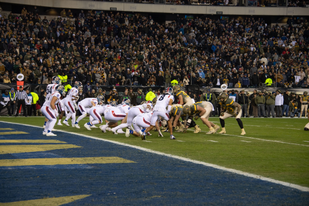 Ein Fußballspiel im Gange auf einem Stadionfeld mit Spielern in Sportbekleidung und Helmen auf dem Boden, Zuschauer und Stadionmerkmale sind im Hintergrund sichtbar.