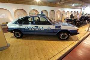 Ein Polizeiwagen in einem Museum ausgestellt, umgeben von Motorrädern und anderen Fahrzeugen, mit einer Säule mit einer Tafel auf der linken Seite und Plakaten an der Wand im Hintergrund, beleuchtet von Lampen auf dem Dach.
