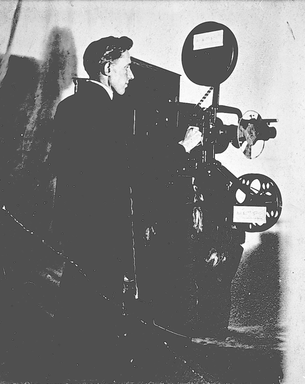 Schwarz-weiß-Foto eines Mannes in einem Anzug, der neben einem Filmprojektor steht und konzentriert aussieht.