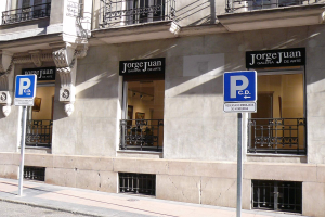 Ein mehrstöckiges Gebäude mit Fenstern, Geländern, Türen und Säulen, das Werbetafeln auf Pfosten entlang des Bürgersteigs und sichtbare Innenobjekte zeigt, die eine belebte städtische Szenerie suggerieren.