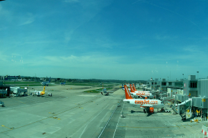 Eine Flughafen-Szene mit Flugzeugen, Gebäuden, Polen, Fahrzeugen und Bäumen, mit einem Himmel darüber und einer Straße darunter und einem Zaun im Vordergrund.