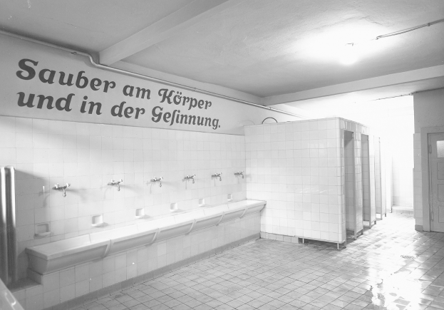 Schwarz-weiß-Foto einer öffentlichen Toilette mit Fliesenboden, Wandtext, mehreren Wasserhähnen, einer Tür und einer Deckenlampe.