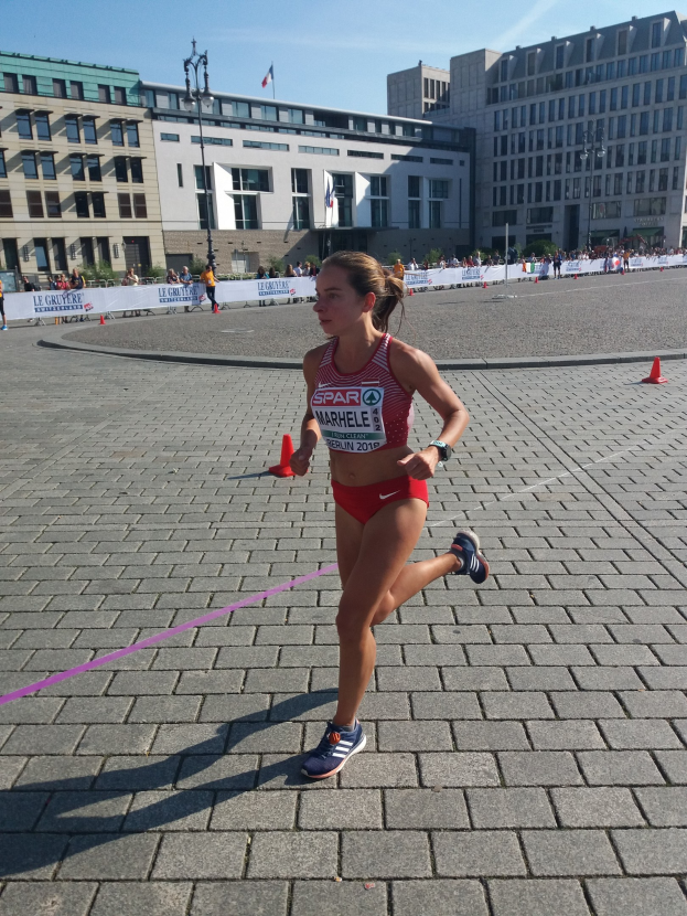 Eine Frau in Sportbekleidung läuft durch eine Marathon-Strecke, die von Verkehrskegeln, einem Band und einer Fahne markiert ist, mit Zuschauern und Straßenelementen unter einem bewölkten Himmel.