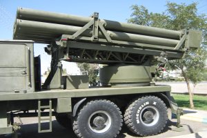 Ein Militärlastwagen mit einem Raketenwerfer fährt auf einer Straße, umgeben von Gras, Bäumen, Gebäuden und einem Maschendrahtzaun, unter einem klaren blauen Himmel.