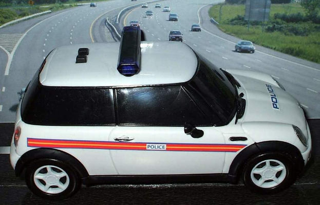 Weißer Polizeiwagen auf einer Straße mit mehreren Fahrzeugen, Gras und Bäumen auf beiden Seiten, aufgenommen außerhalb einer Stadt.