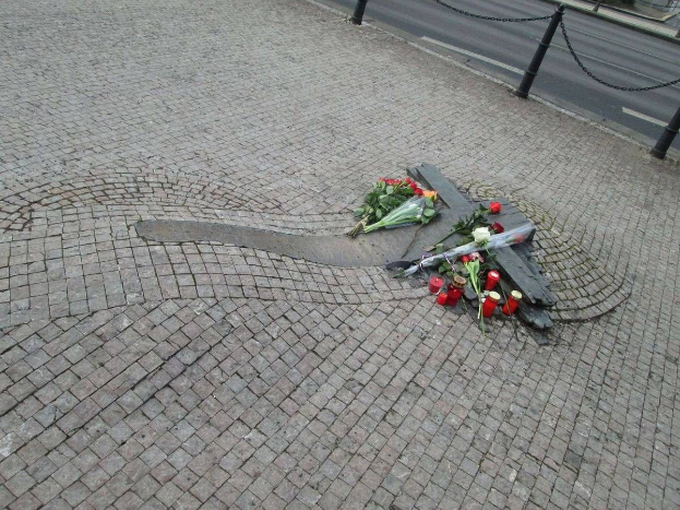 Gedenken mit Blumen, Kerzen und verstreuten Buketts am Straßenrand, umgeben von Bäumen, Gebäuden und Fahrzeugen mit Kettenpfählen.