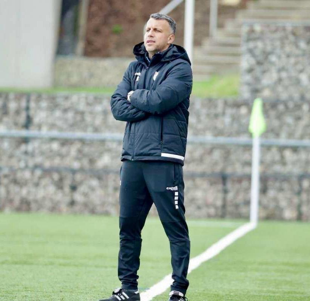 Ein Fußballtrainer auf einem Feld mit verschränkten Armen, der eine schwarze Jacke und Hose trägt, mit einem Zaun, Treppe und Wand im Hintergrund.